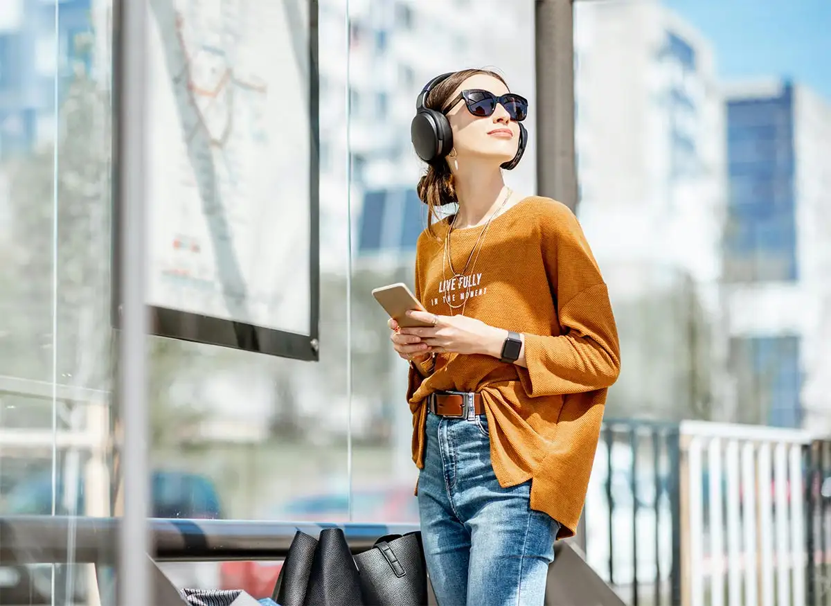 a happy woman holding a mobile phone standing in the bus drop and pick up station